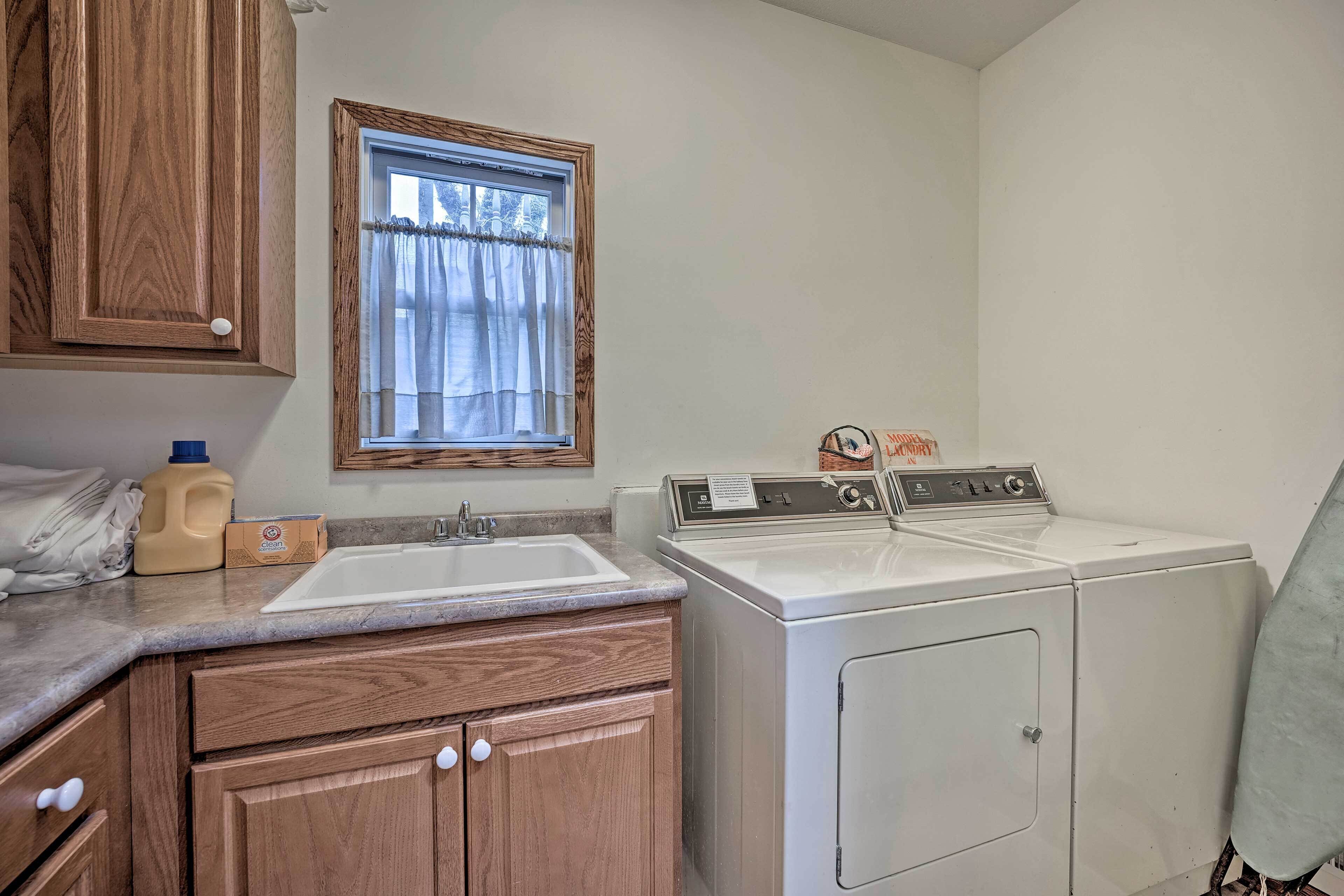 A laundry room with a sink, two washing machines, wooden cabinets, and a window with curtains.