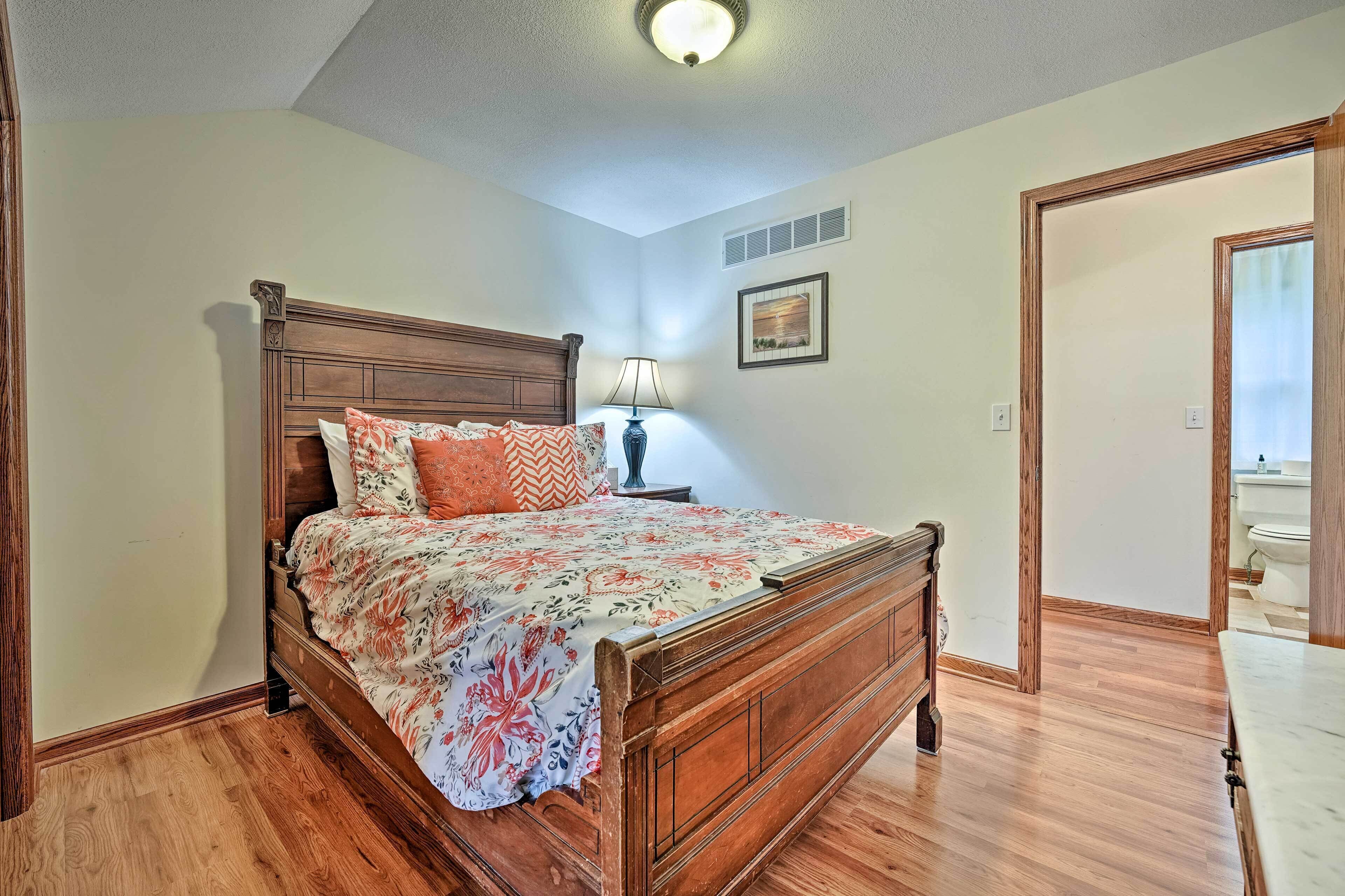 A bedroom with a wooden bed, floral bedding, a nightstand with a lamp, and a bathroom visible through an open door.
