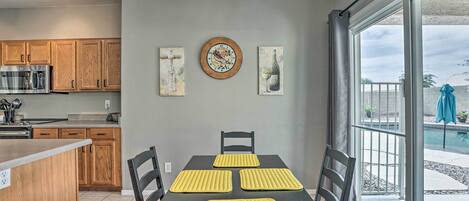 A modern kitchen with wooden cabinets, a stainless steel refrigerator, and a dining area with a table and chairs. There are decorative wall art pieces and a clock on the wall.
