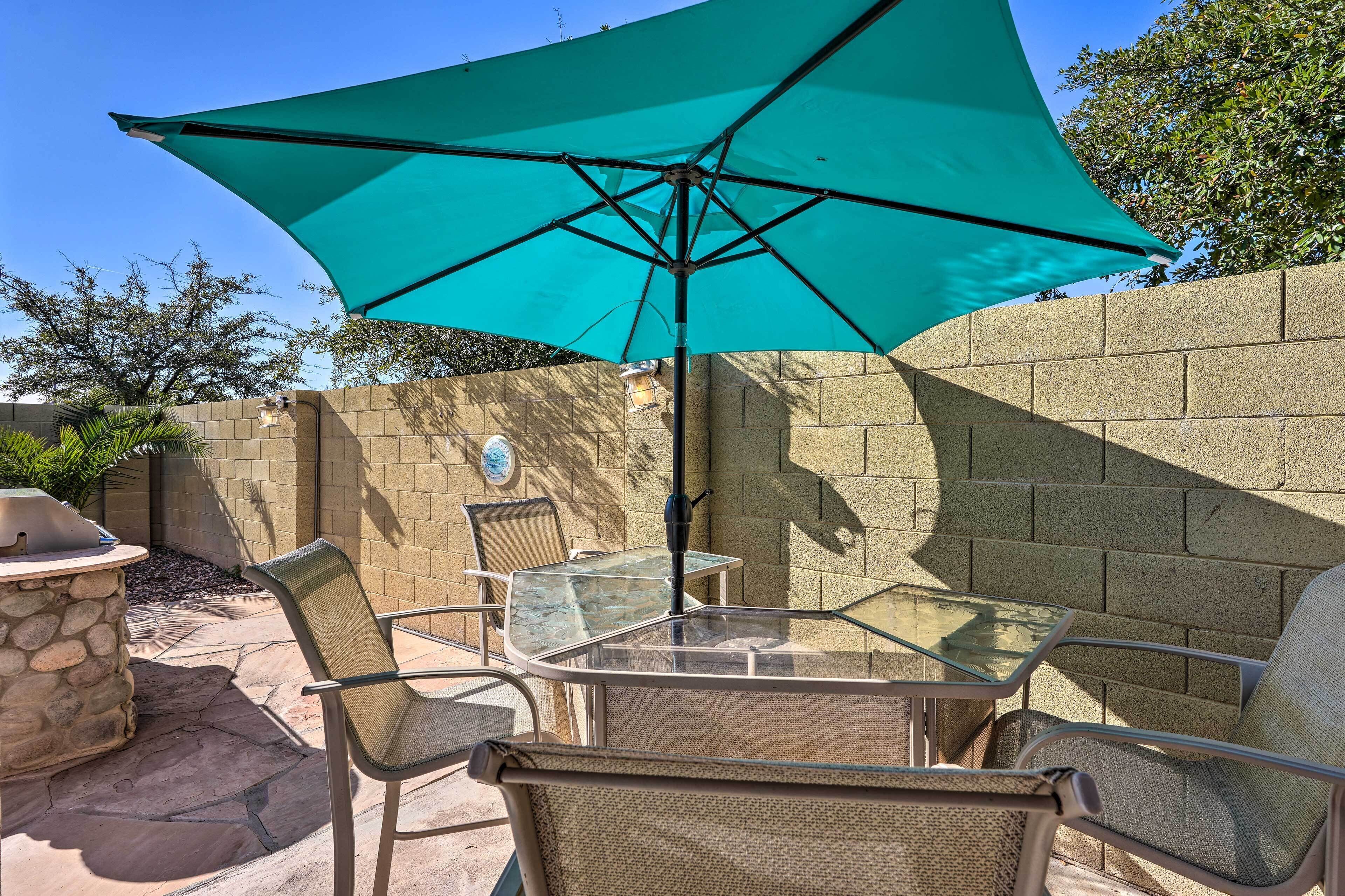 Un patio con una mesa y sillas de vidrio, un parasol azul turquesa y una estación de barbacoa de piedra.
