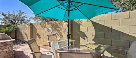 Un patio con una mesa y sillas de vidrio, un parasol azul turquesa y una estación de barbacoa de piedra.