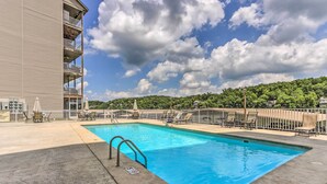 A rectangular swimming pool with a metal ladder, surrounded by lounge chairs and umbrellas, with a building and a forested hill in the background. - Lake & Poolside Fun! Water-view Camdenton Condo (Camdenton)