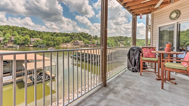 A covered outdoor area with a wooden deck, a table and chairs, a ceiling fan, and a view of a lake with boats and houses in the background. - Lake & Poolside Fun! Water-view Camdenton Condo (Camdenton)