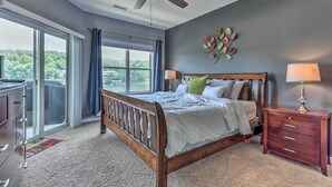 A bedroom with a wooden bed, a ceiling fan, a nightstand, and a view of the outdoors through a large window. - Lake & Poolside Fun! Water-view Camdenton Condo (Camdenton)