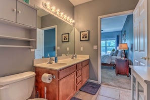 A bathroom with a double sink vanity, a toilet, and a large mirror with lights. There is a door leading to a bedroom with a bed and a nightstand. - Lake & Poolside Fun! Water-view Camdenton Condo (Camdenton)