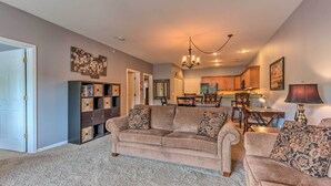A living room with a beige sofa set, a coffee table, and a bookshelf. There is a dining area with a table and chairs in the background. - Lake & Poolside Fun! Water-view Camdenton Condo (Camdenton)