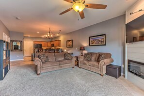 A living room with a beige sofa set, a ceiling fan, a fireplace, and a kitchen in the background. - Lake & Poolside Fun! Water-view Camdenton Condo (Camdenton)