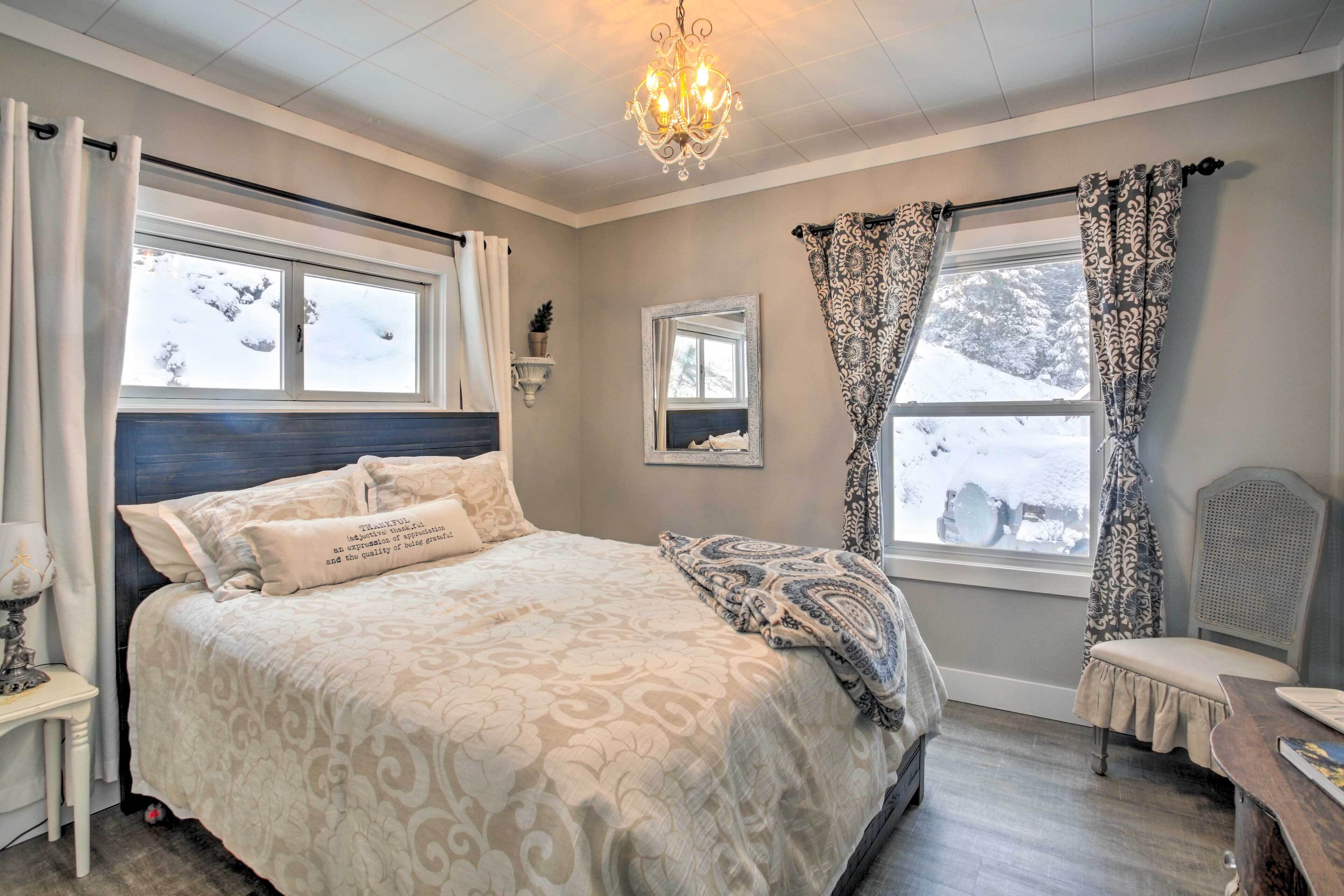 Une chambre à coucher avec un grand lit, deux fenêtres avec des rideaux, un lustre, une table de chevet et une chaise.
