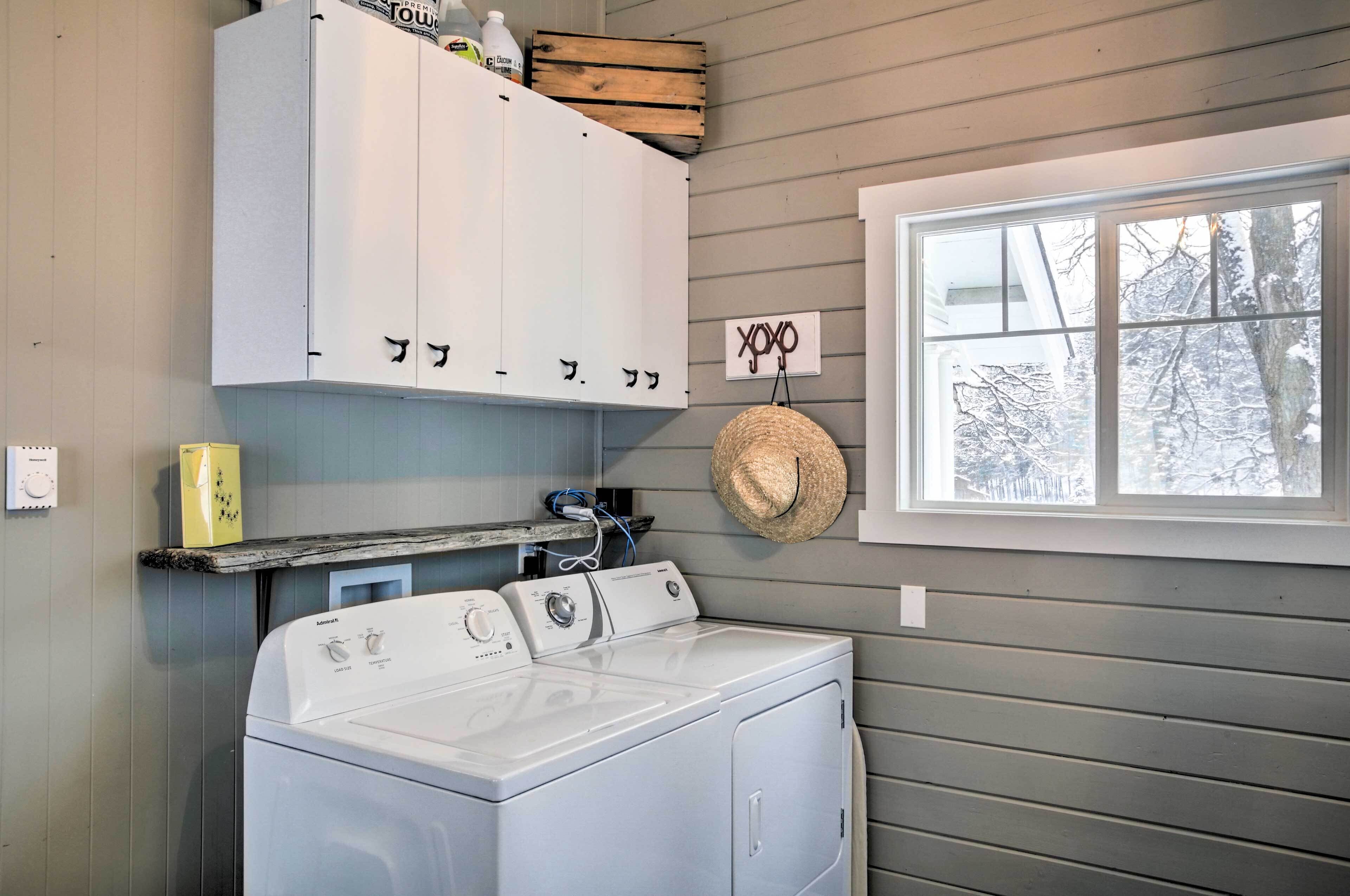 A laundry area with a washer and dryer, wooden cabinets, a straw hat, and a window with a view of snow-covered trees.