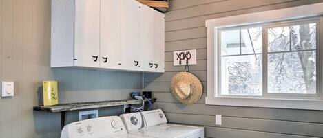 A laundry area with a washer and dryer, wooden cabinets, a straw hat, and a window with a view of snow-covered trees.