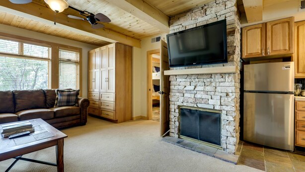 A cozy living room with a stone fireplace, wooden ceiling beams, a flat-screen TV mounted above it, and a modern stainless steel refrigerator. - Ski-in/ski-out Studio in Aspen Snowmass Ski Resort (Snowmass Village)