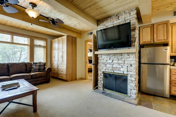 A cozy living room with a stone fireplace, wooden ceiling beams, a flat-screen TV mounted above it, and a modern stainless steel refrigerator. - Ski-in/ski-out Studio in Aspen Snowmass Ski Resort (Snowmass Village)