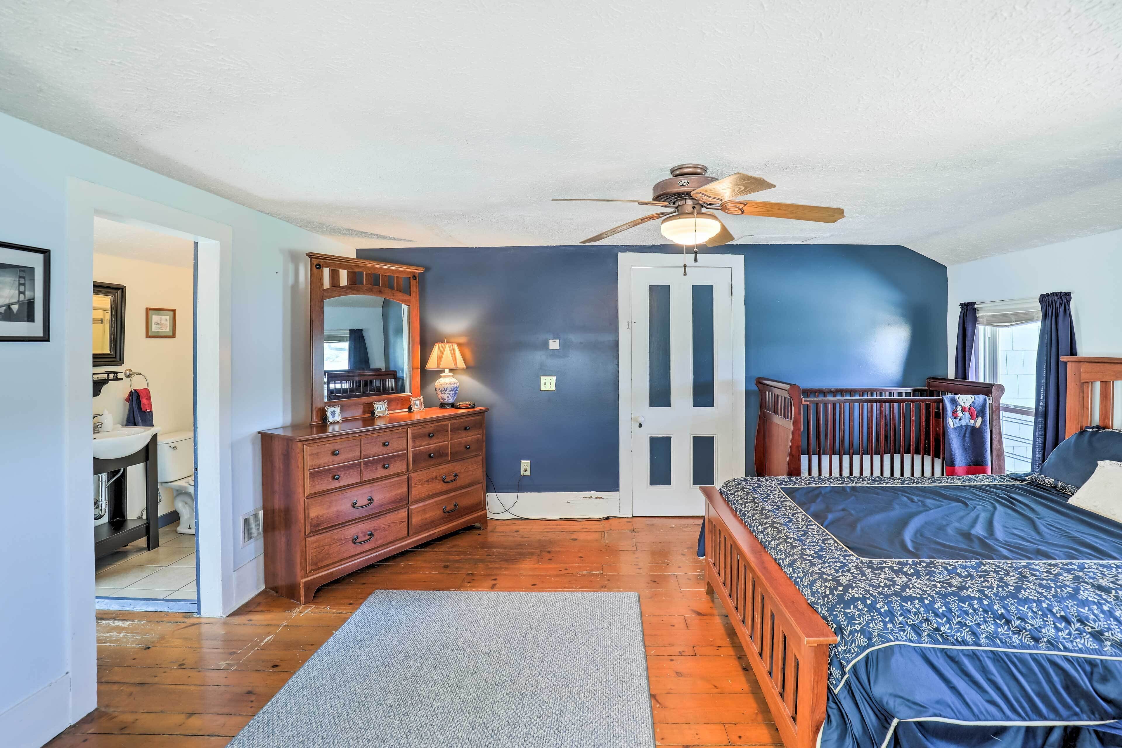 A bedroom with a bed, dresser, mirror, ceiling fan, and a door leading to another room.