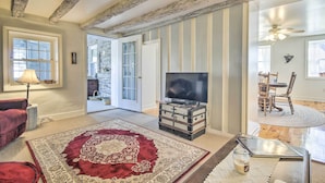 Un salon avec un téléviseur, un tapis rouge, une commode en bois et un coin repas visible à travers une porte ouverte.
