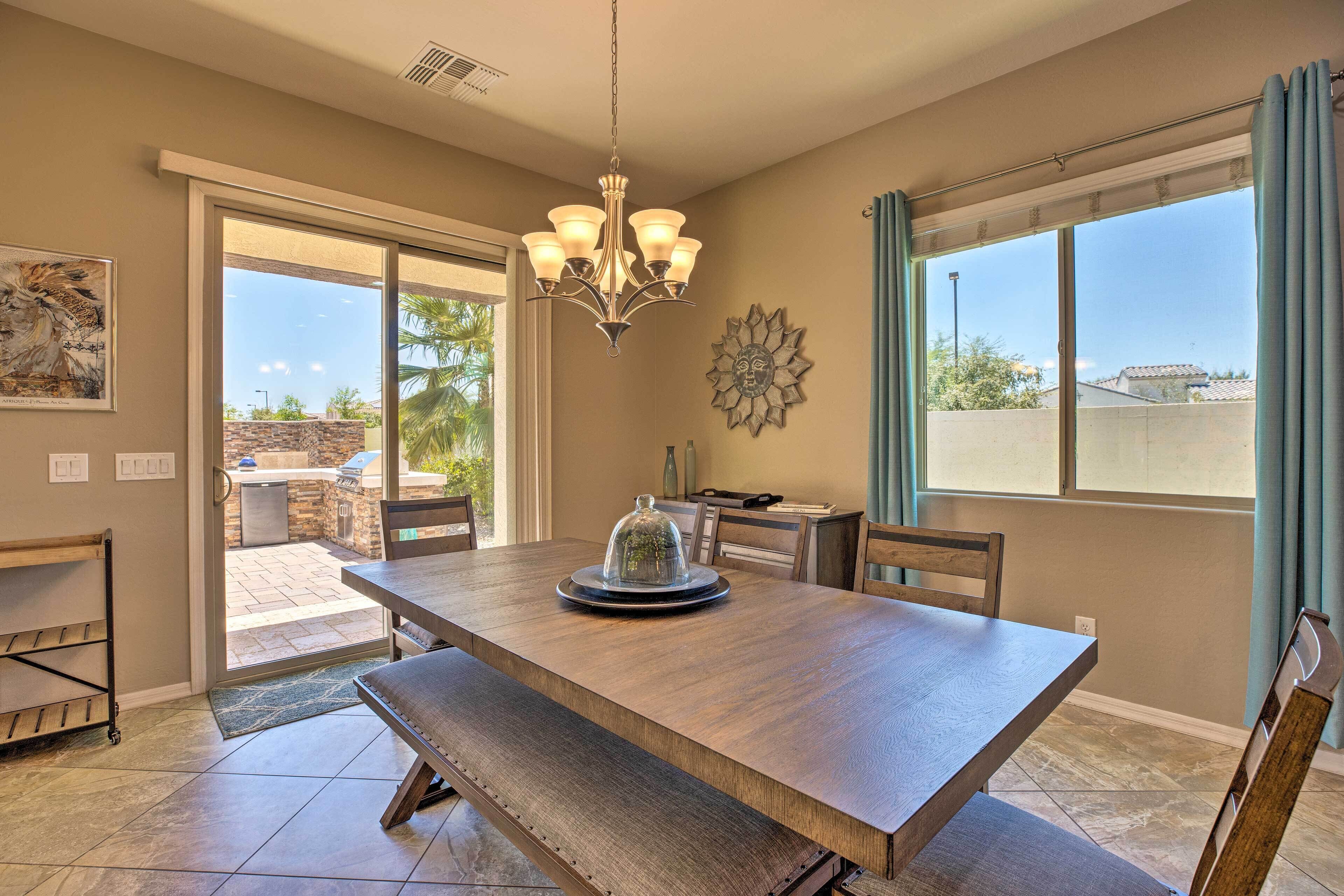 A dining area with a large wooden table, bench seating, a chandelier, and a view through a sliding glass door to an outdoor patio.
