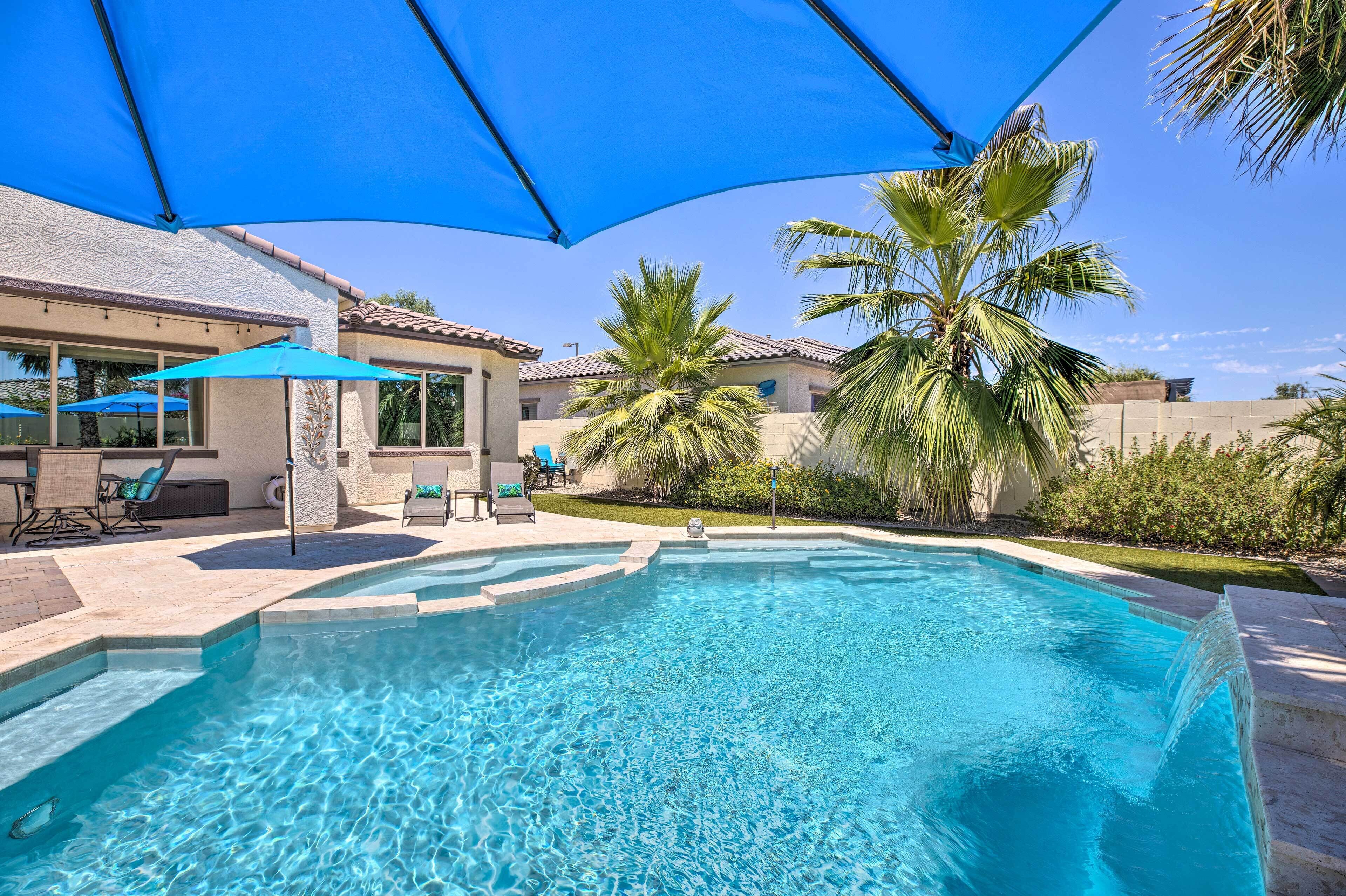 A swimming pool with a clear blue water, surrounded by palm trees and a covered patio area with chairs and umbrellas.