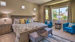 A hotel room with a large bed, two bedside tables with lamps, a bench, a blue armchair, and a view of palm trees through the window. - Modern Azure Home w/ Beautiful Patio, Pool & Spa! (Goodyear)