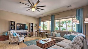 A spacious living room with a large sofa, a coffee table, and a television. There is a ceiling fan, a dining table with chairs, and a bookshelf. - Modern Azure Home w/ Beautiful Patio, Pool & Spa! (Goodyear)