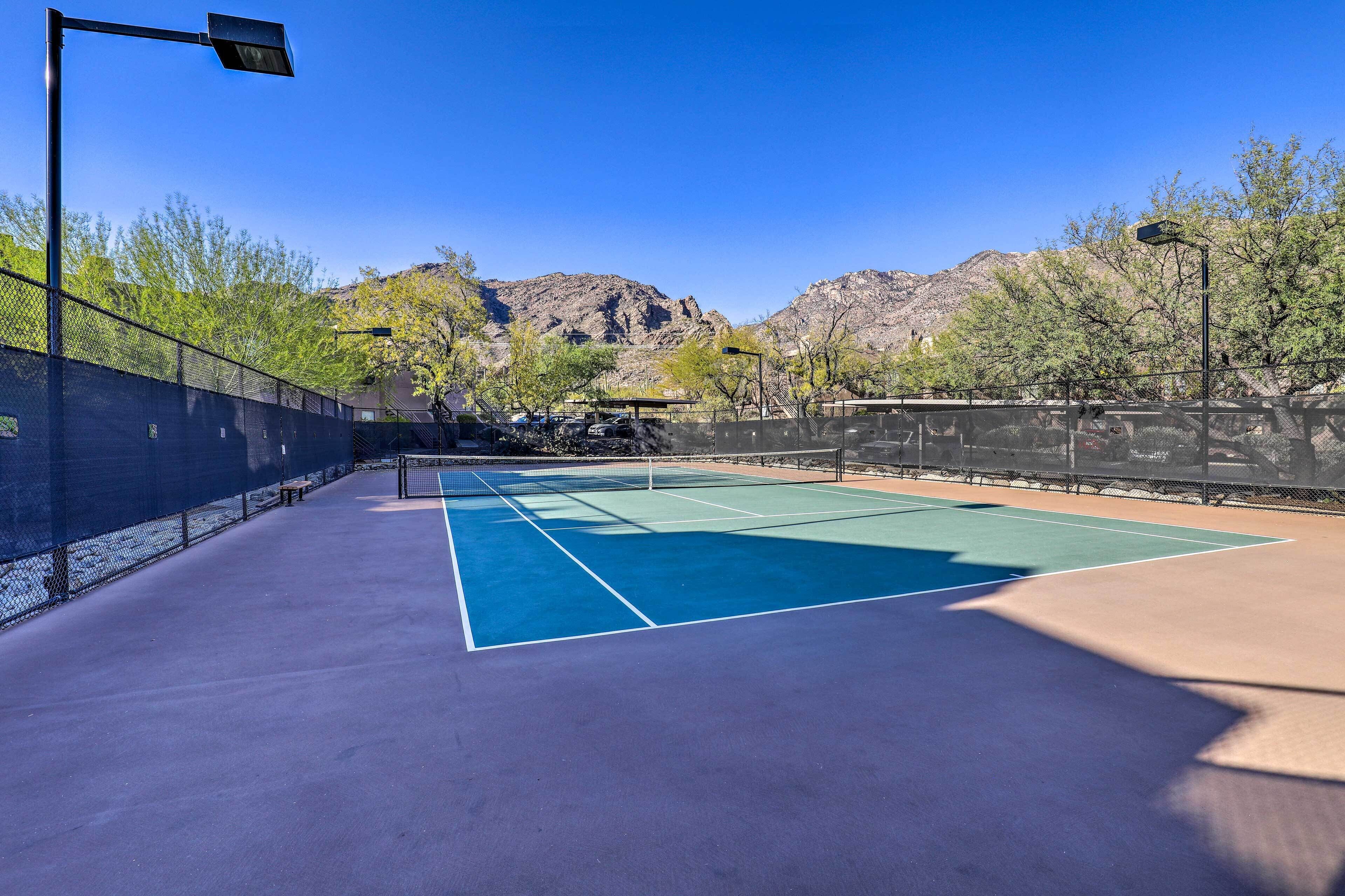 An outdoor tennis court with a net, surrounded by a fence, and mountains in the background.