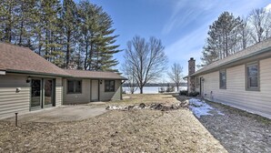 A two-story house with a stone chimney, a covered patio, and a view of a lake. - Lakefront Escape w/ Fishing Pier & Snowmobiling! (Park Falls)