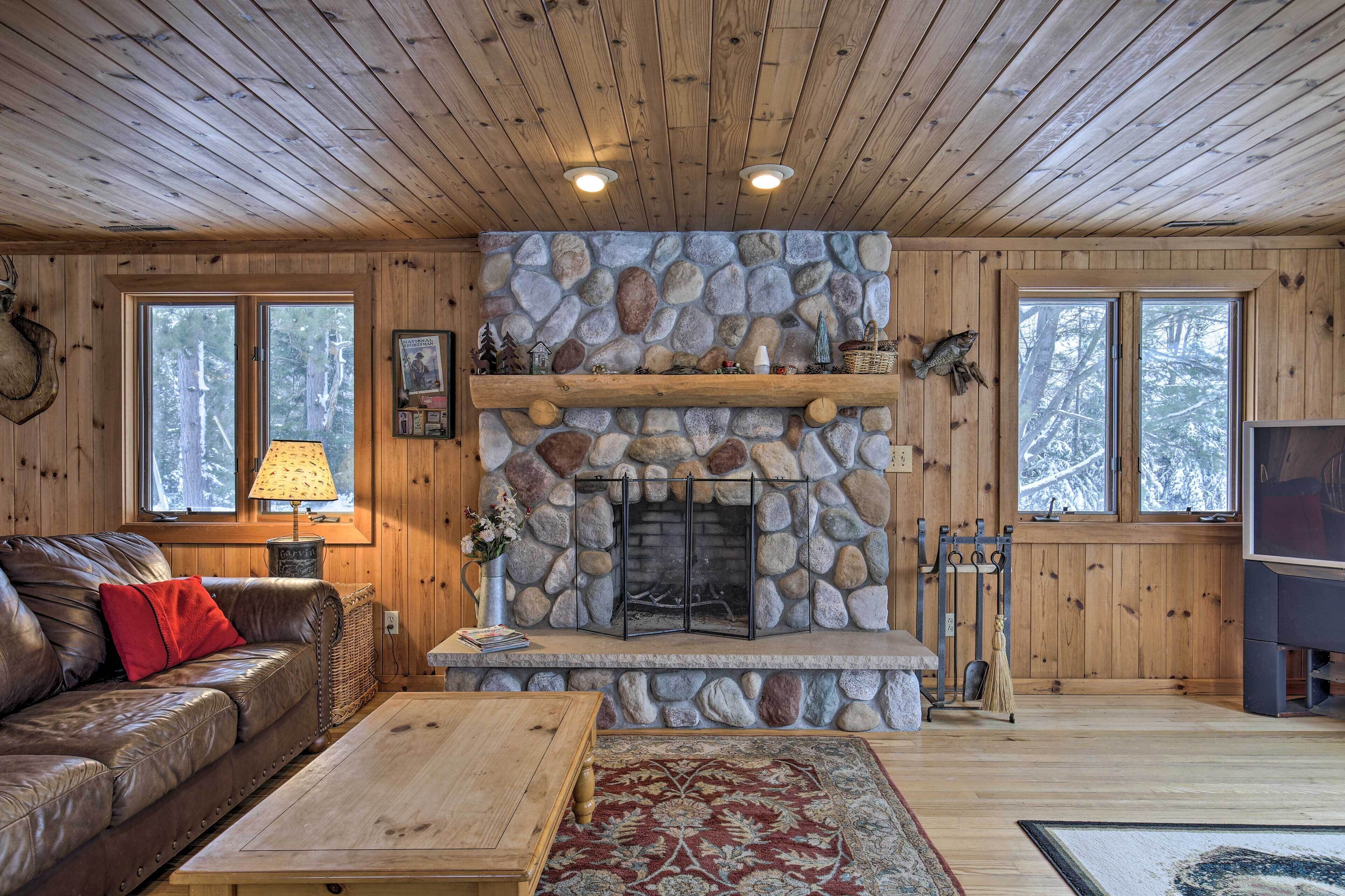 A cozy cabin interior with a stone fireplace, wooden walls, a leather sofa, and a large window.