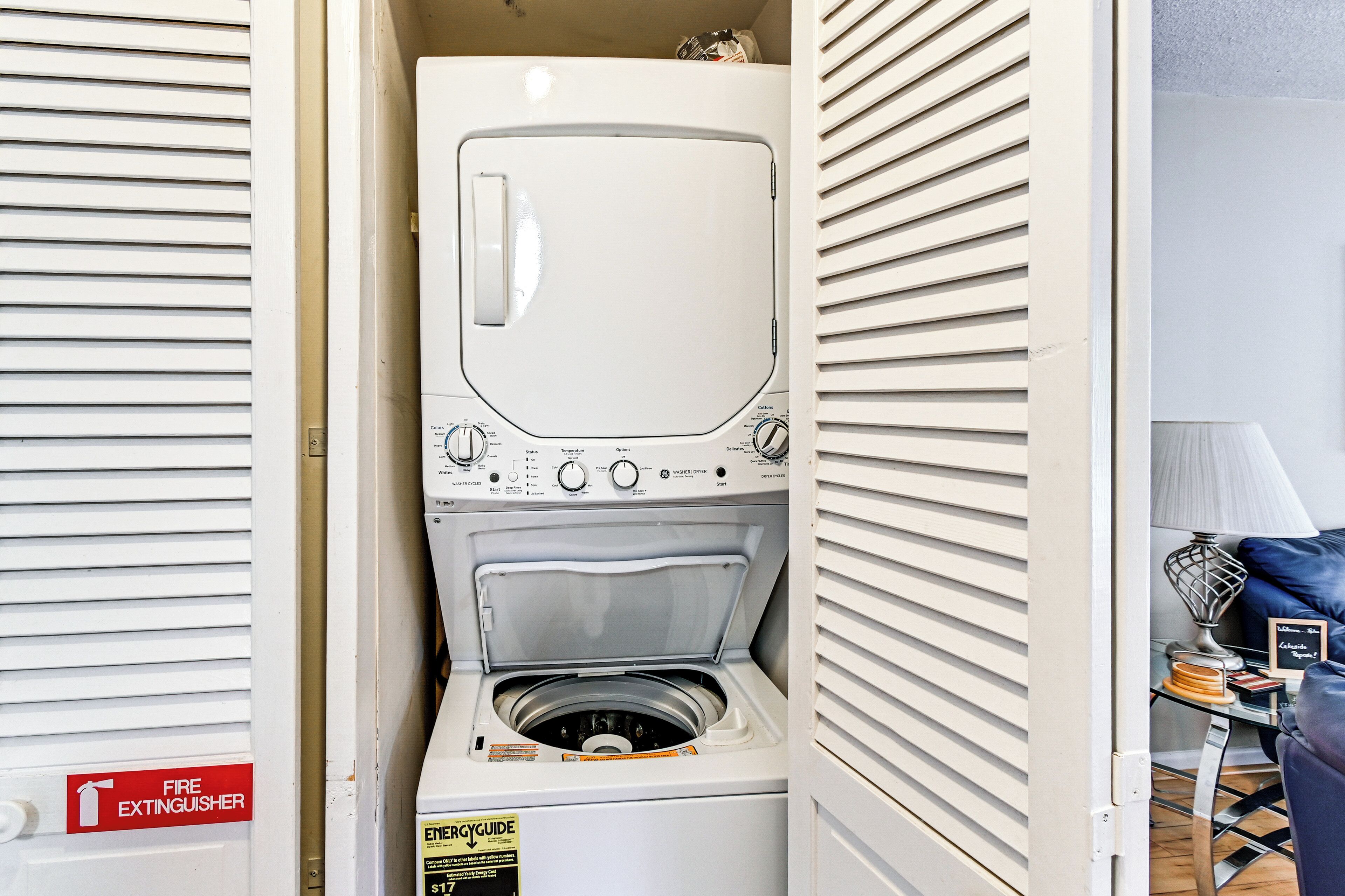 A compact, white laundry room with a built-in washer and dryer, a fire extinguisher, and a small table with a lamp and decorative items.