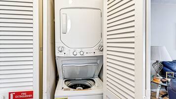 A compact, white laundry room with a built-in washer and dryer, a fire extinguisher, and a small table with a lamp and decorative items.