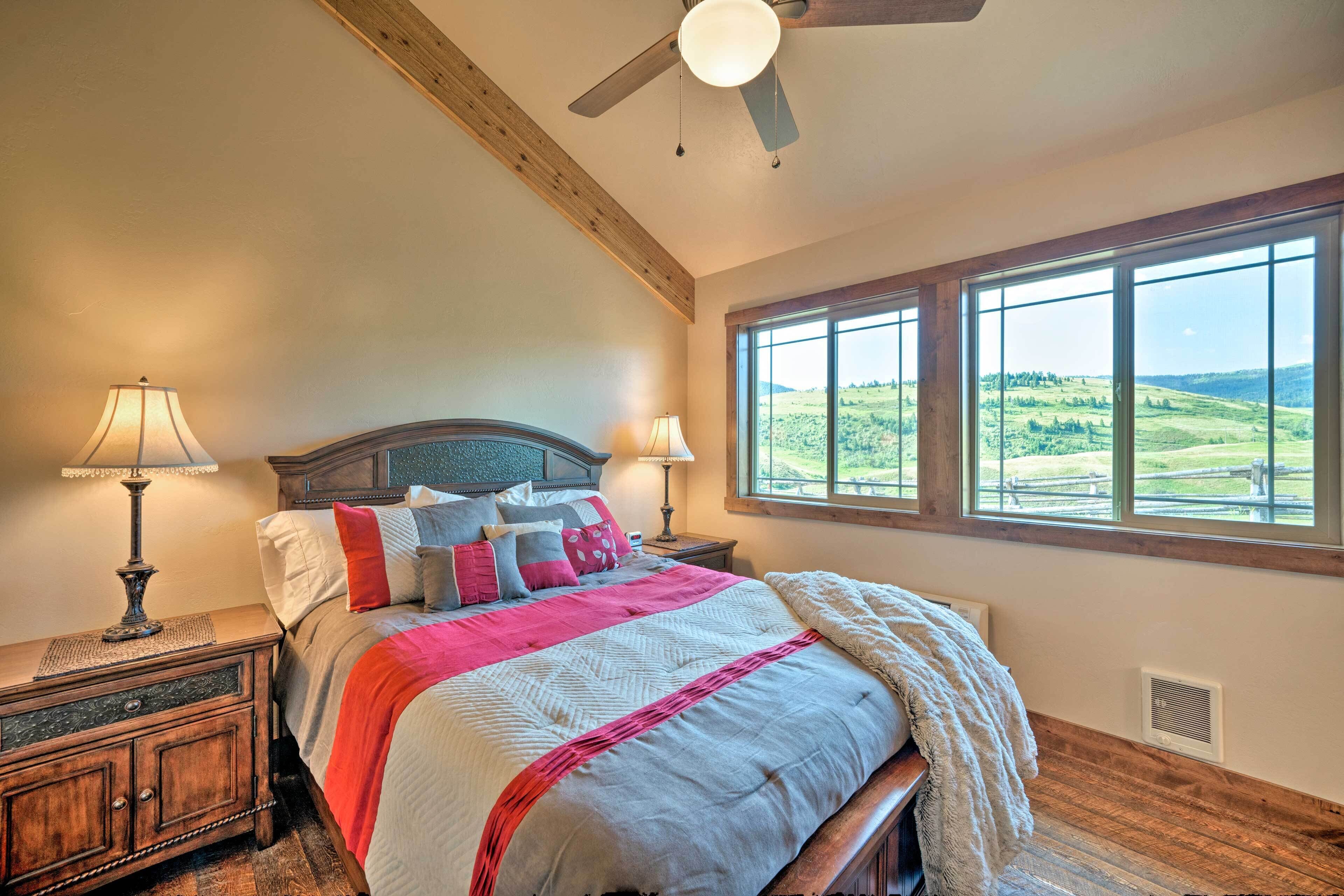 A bedroom with a large bed, two bedside lamps, a ceiling fan, and a view of a green landscape through large windows.