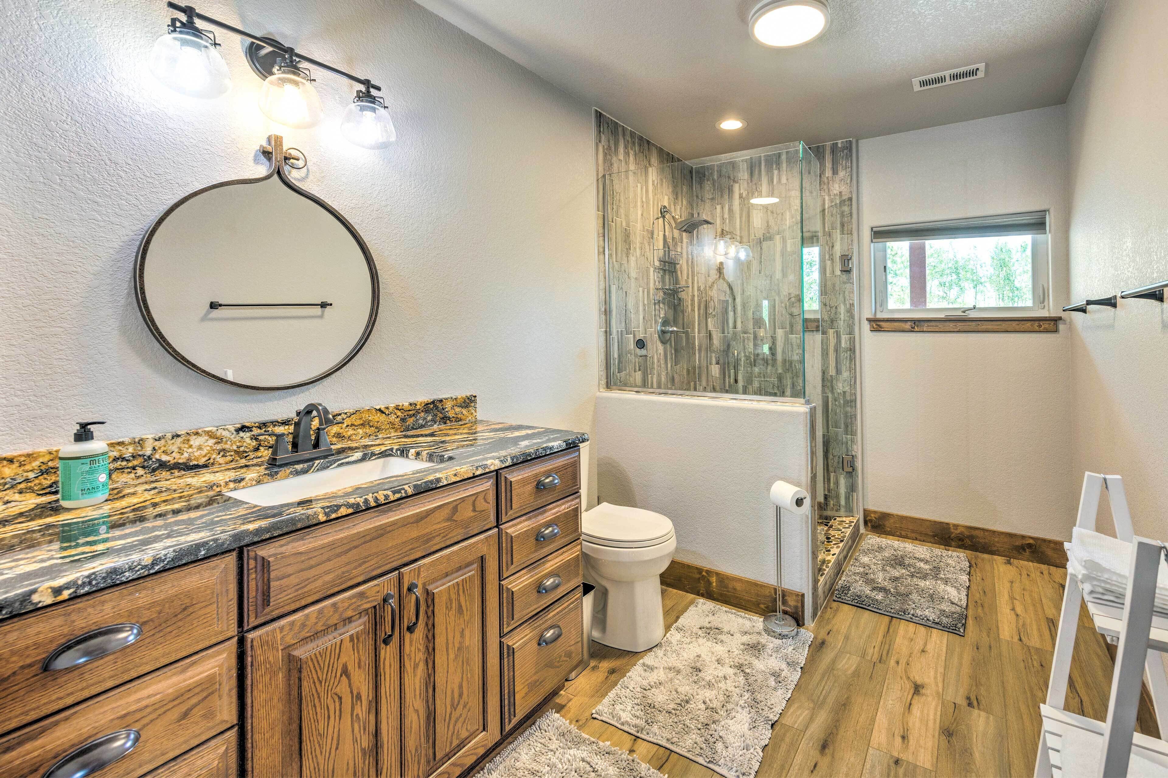 A bathroom with wooden cabinets, a granite countertop, a round mirror, a toilet, and a glass shower enclosure.