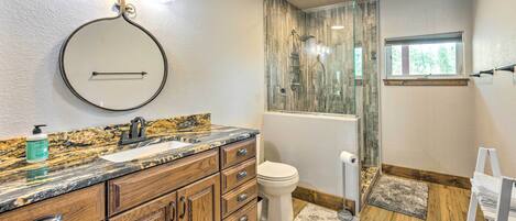A bathroom with wooden cabinets, a granite countertop, a round mirror, a toilet, and a glass shower enclosure.
