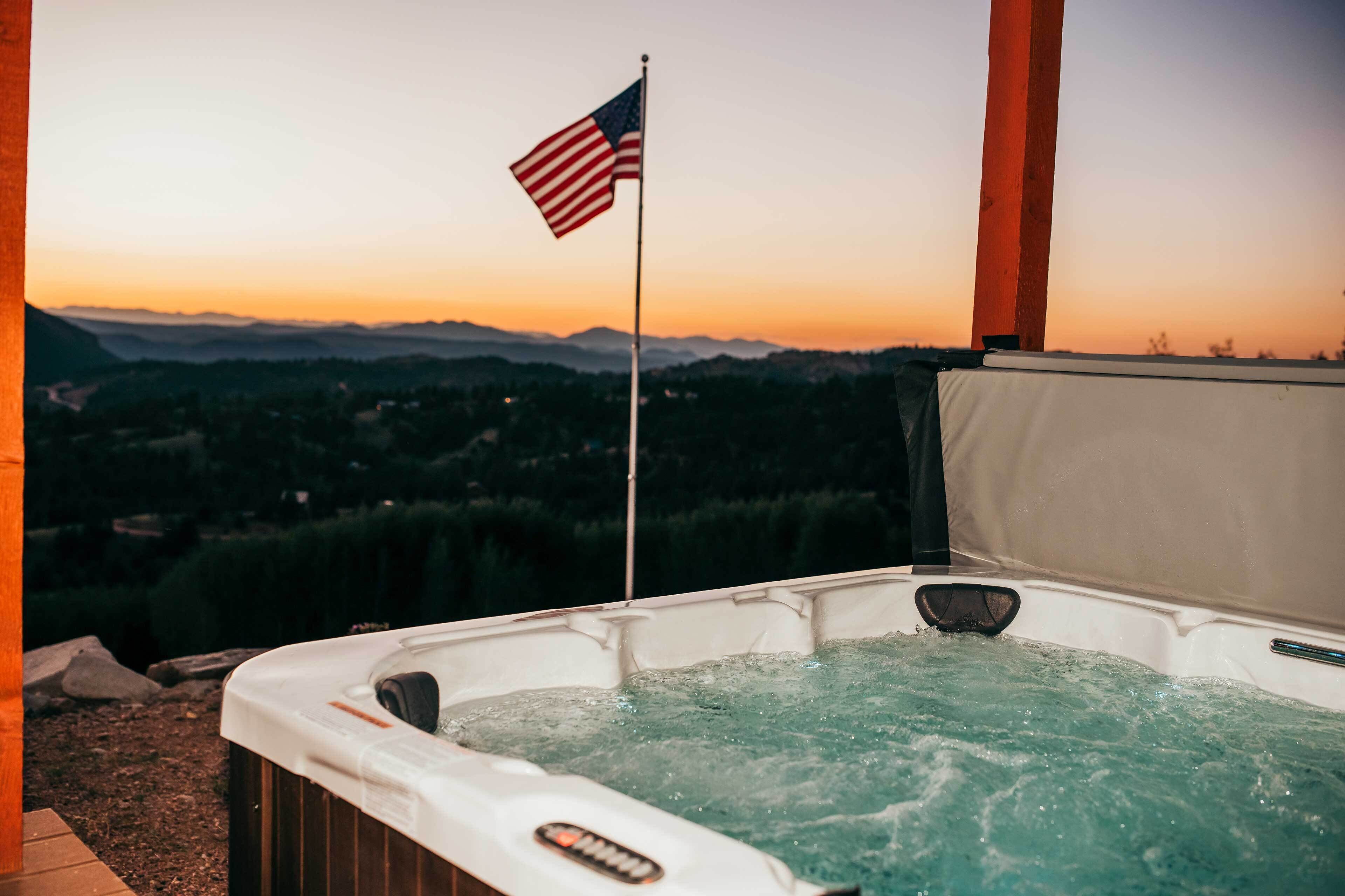 Uma banheira de hidromassagem com uma bandeira americana, rodeada por um deck de madeira e com vista para uma paisagem montanhosa.