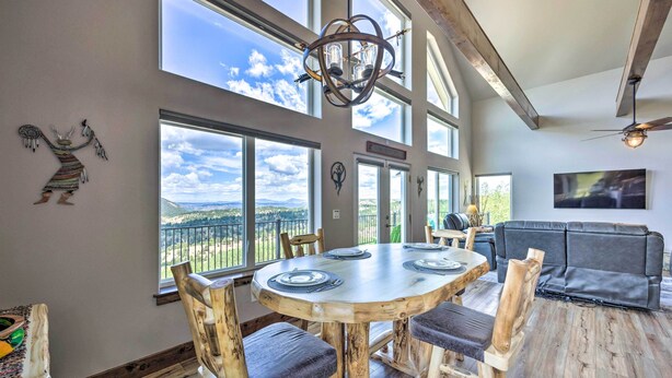 A spacious dining area with a large wooden table and chairs, large windows with a view of the outdoors, and a modern chandelier. - Hot Tub & Mtn View! Cripple Creek Retreat (Cripple Creek)