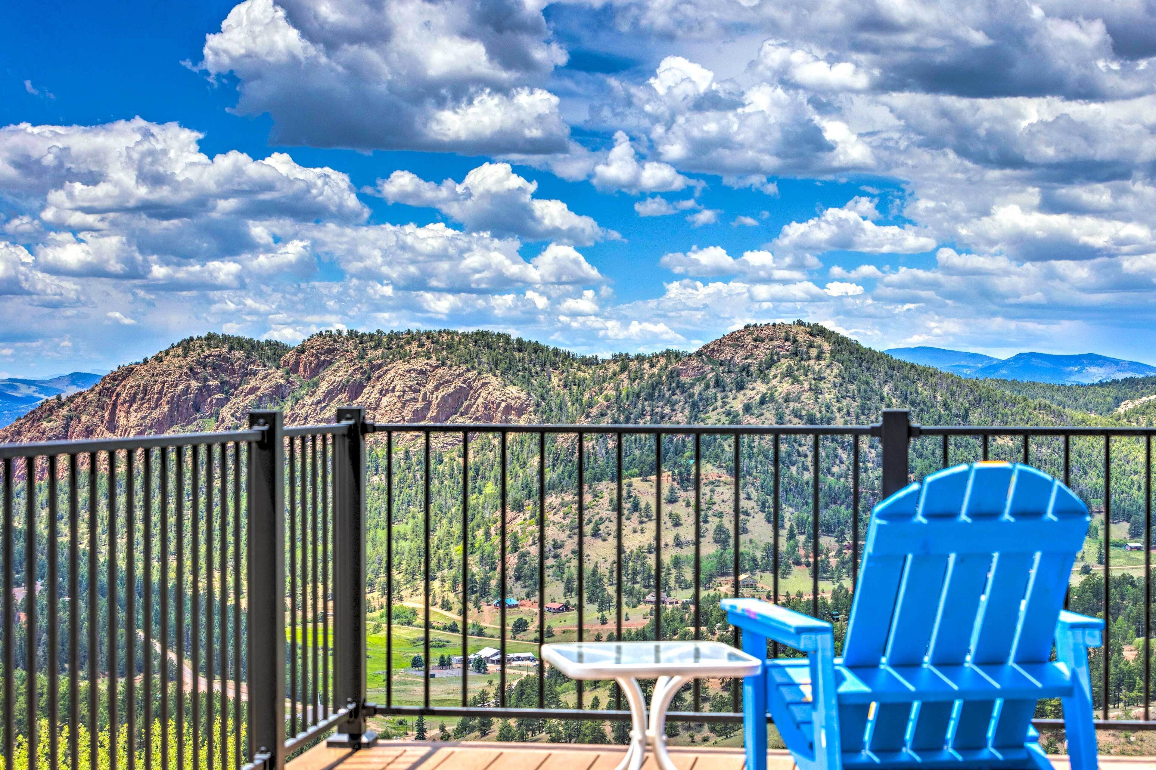 Un balcón con una silla Adirondack azul y una mesa pequeña, con vistas a un paisaje montañoso y cielo despejado.