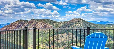 Un balcón con una silla Adirondack azul y una mesa pequeña, con vistas a un paisaje montañoso y cielo despejado.