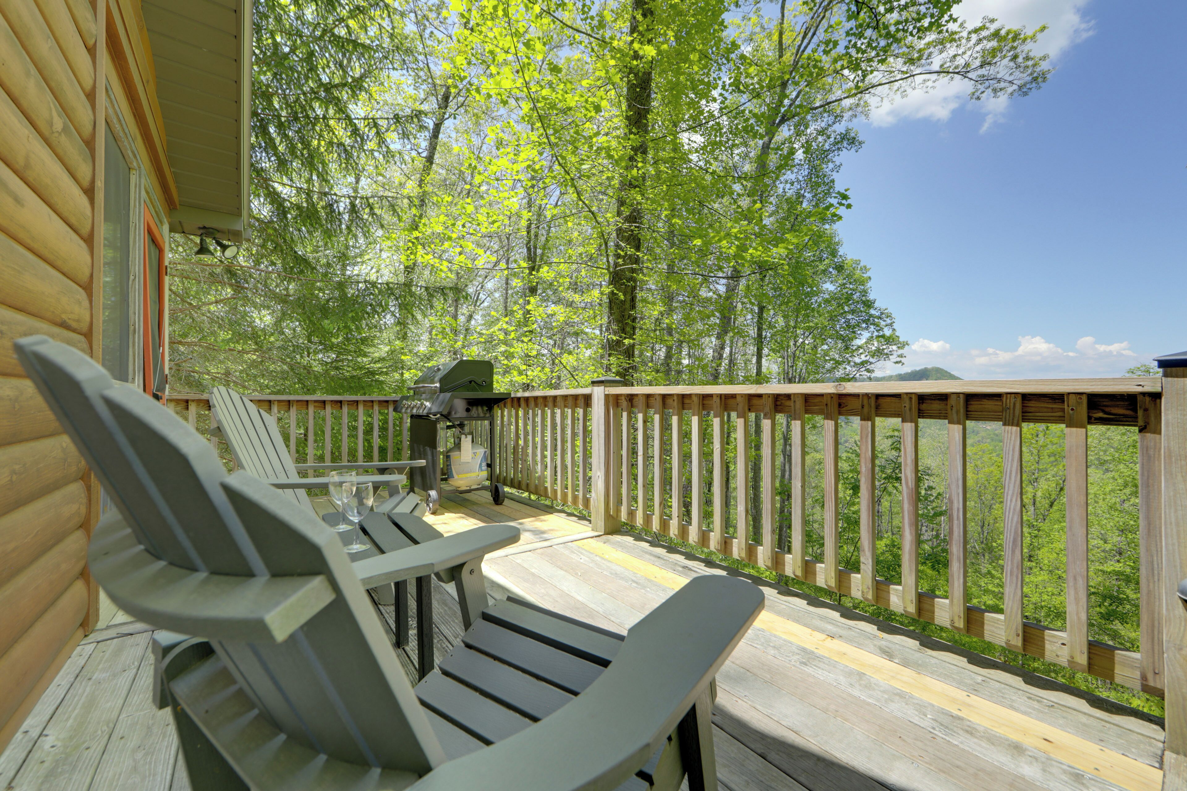 A wooden deck with two Adirondack chairs and a grill, overlooking a forested area.