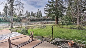 A riverside view with a wooden deck, a grassy area, and a row of houses in the background. - Hot Tub & Views: Spacious Getaway on Rogue River (Grants Pass)