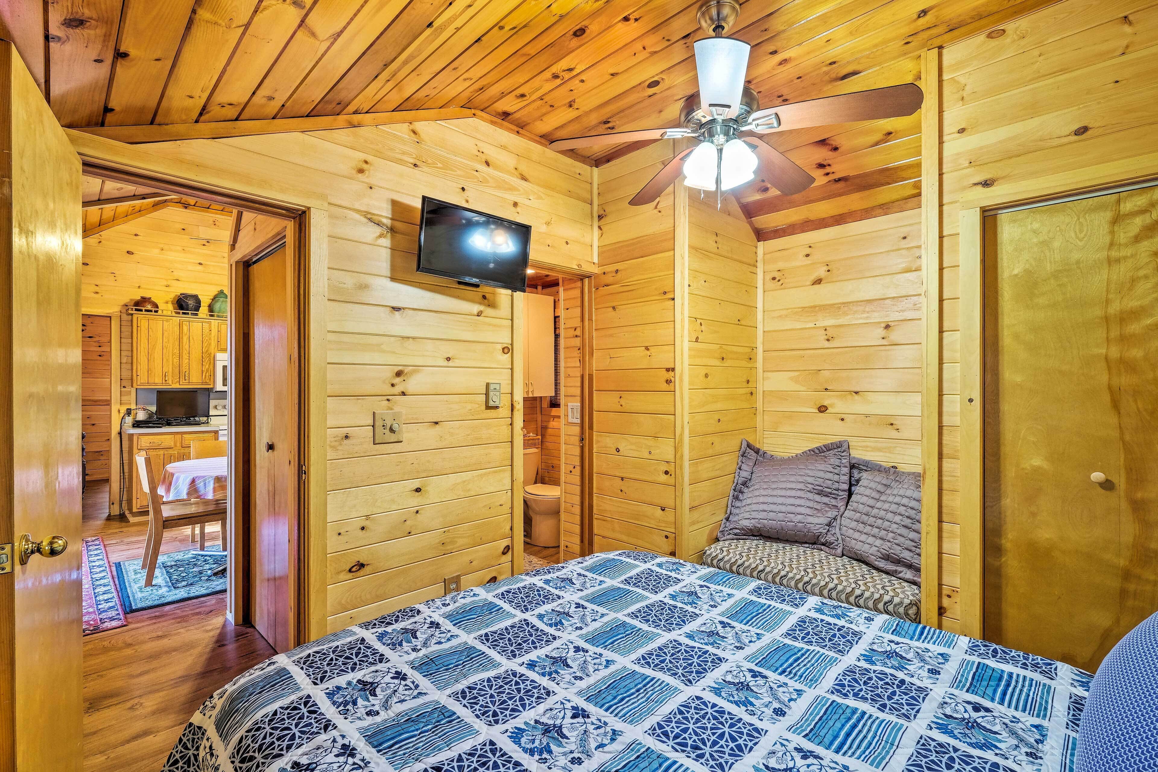A wooden cabin room with a bed, a ceiling fan, a television, and a small kitchen area visible through an open door.