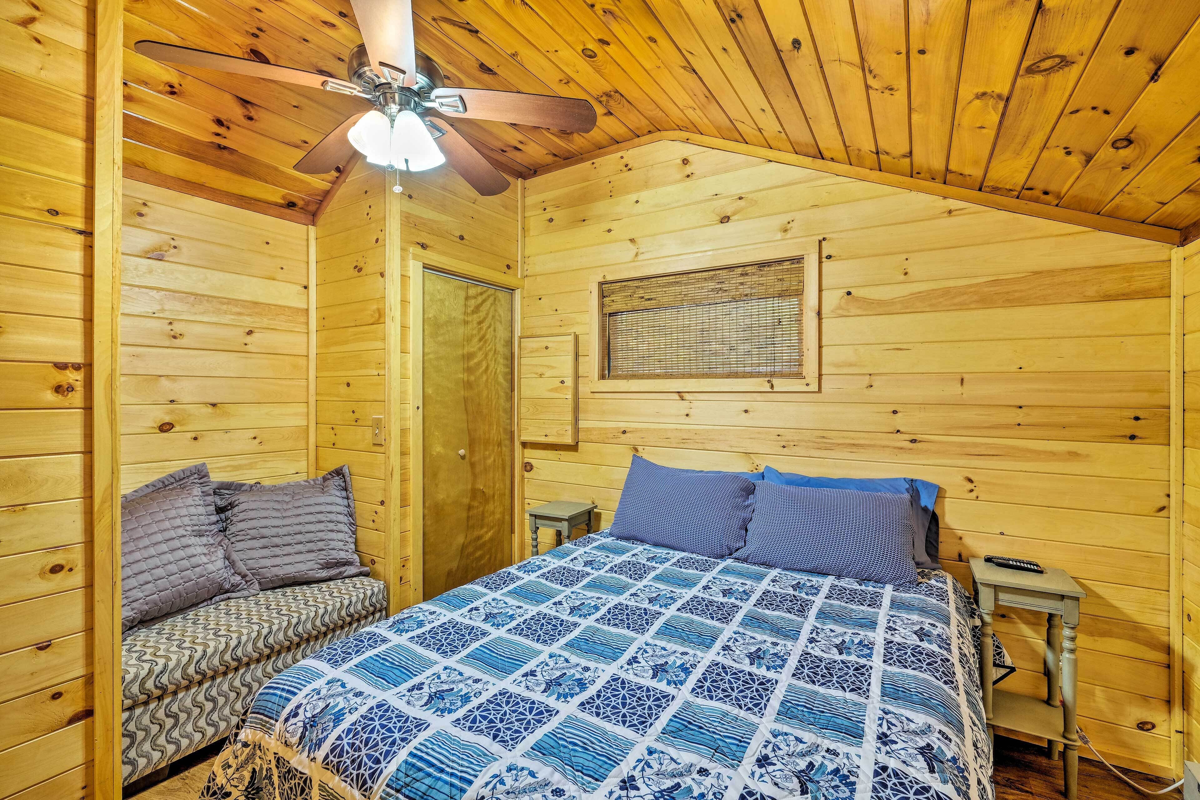 Um quarto de cabana de madeira aconchegante, com uma cama, um banco, uma mesa pequena e um ventilador de teto.
