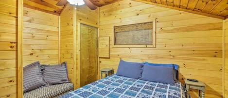 Um quarto de cabana de madeira aconchegante, com uma cama, um banco, uma mesa pequena e um ventilador de teto.