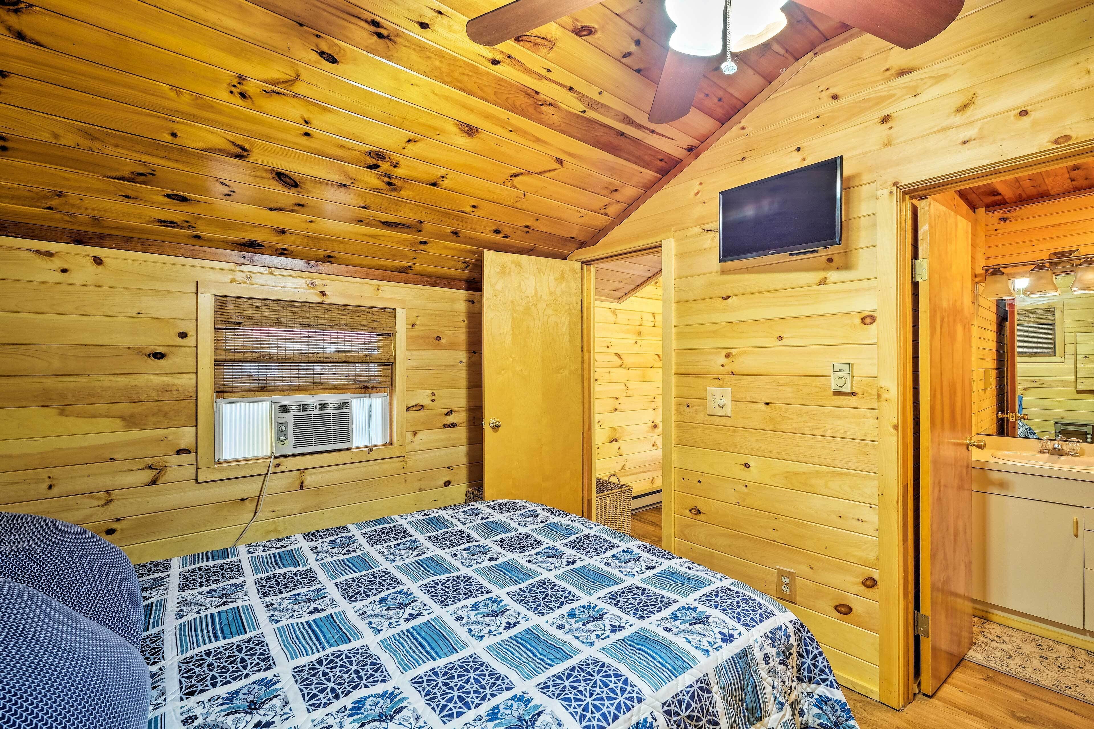 Habitación de cabaña de madera con una cama, televisor, ventana con persianas y baño con lavamanos y espejo.