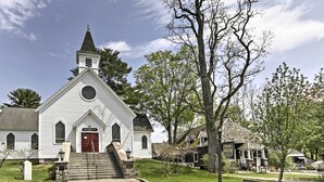 A white church with a red door and a bell tower, surrounded by trees and a bench. - Walk to Beach! Bolton Landing Cottage w/ Deck (Bolton Landing)