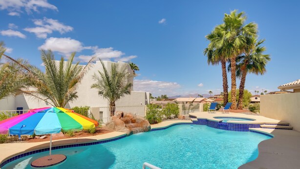 A swimming pool with a curved edge, surrounded by palm trees and a colorful umbrella. - 3 Mi to Lake Havasu: Luxe Home w/ Pool & Spa! (Lake Havasu City)