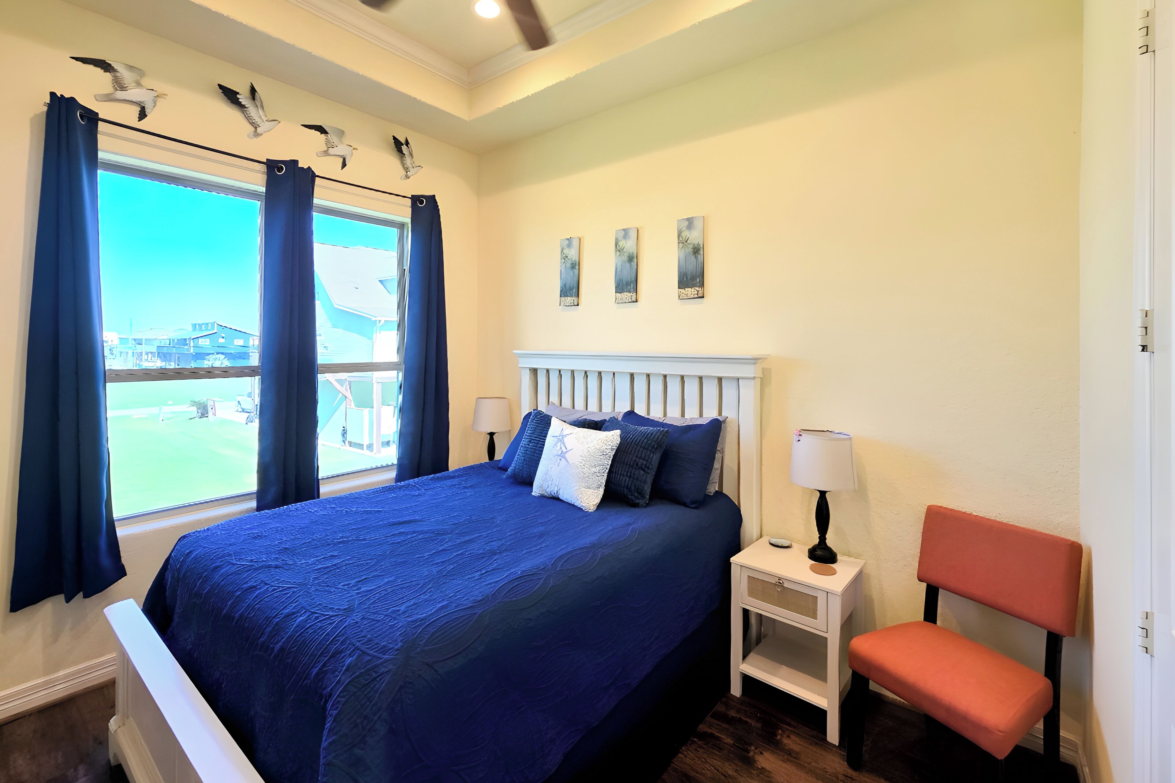 A bedroom with a large bed, a window with curtains, a nightstand, a chair, and artwork on the wall.