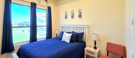 A bedroom with a large bed, a window with curtains, a nightstand, a chair, and artwork on the wall.