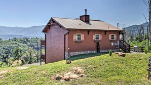 A wooden cabin with a stone foundation, a chimney, and a small porch. It is situated on a grassy hill with a view of a forested mountain range.