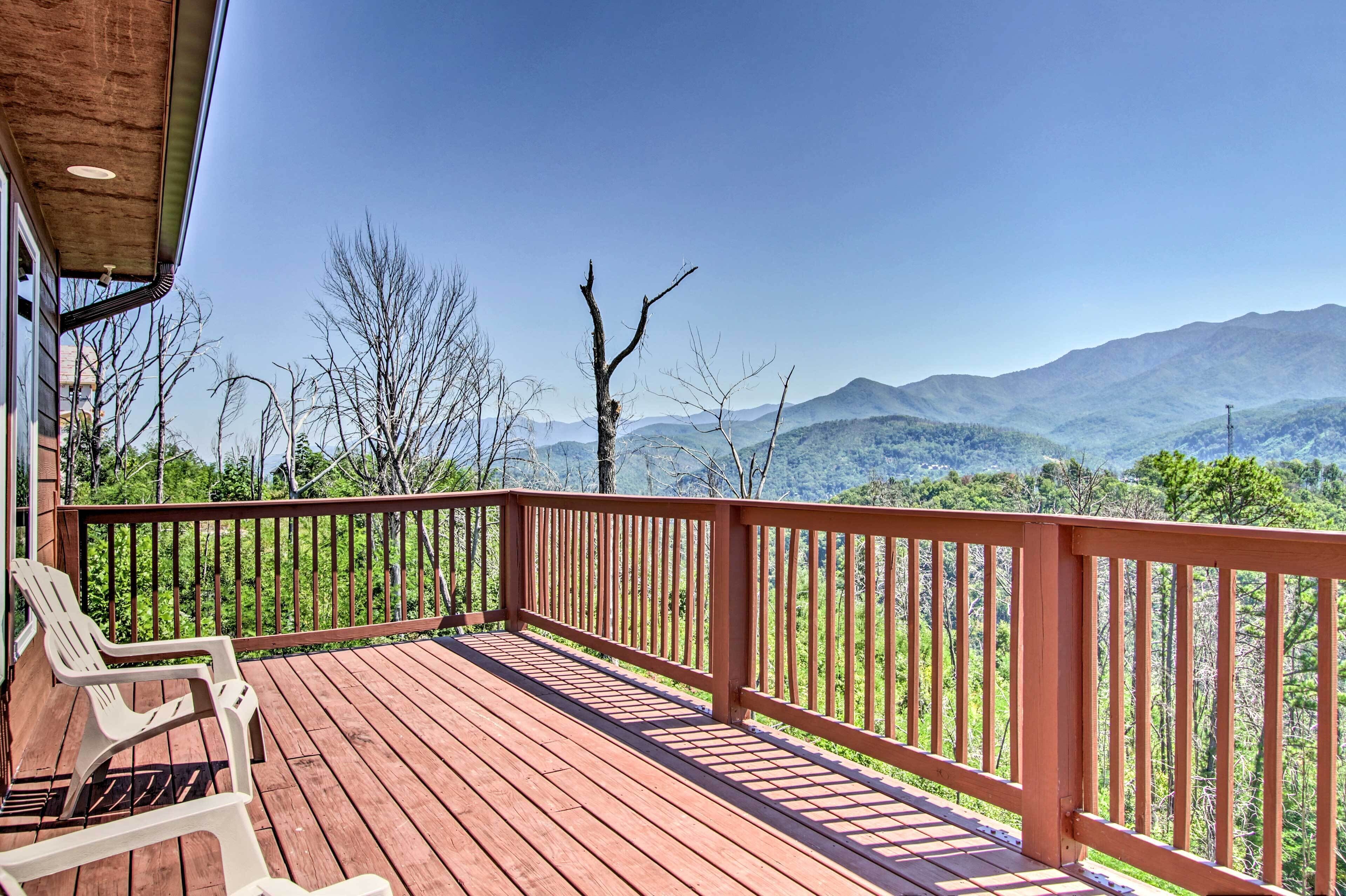 A wooden deck with a railing, two white chairs, and a view of mountains.