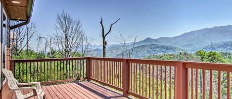A wooden deck with a railing, two white chairs, and a view of mountains.