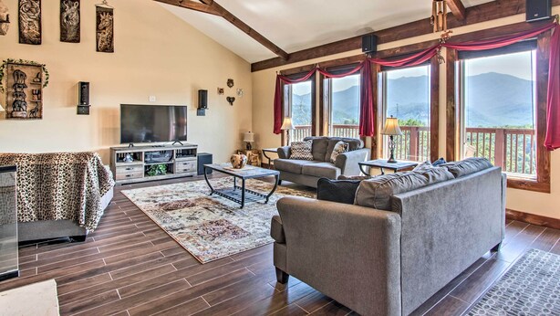 A spacious living room with a large sofa, a television, and a fireplace. There are large windows with curtains, and a wooden ceiling.