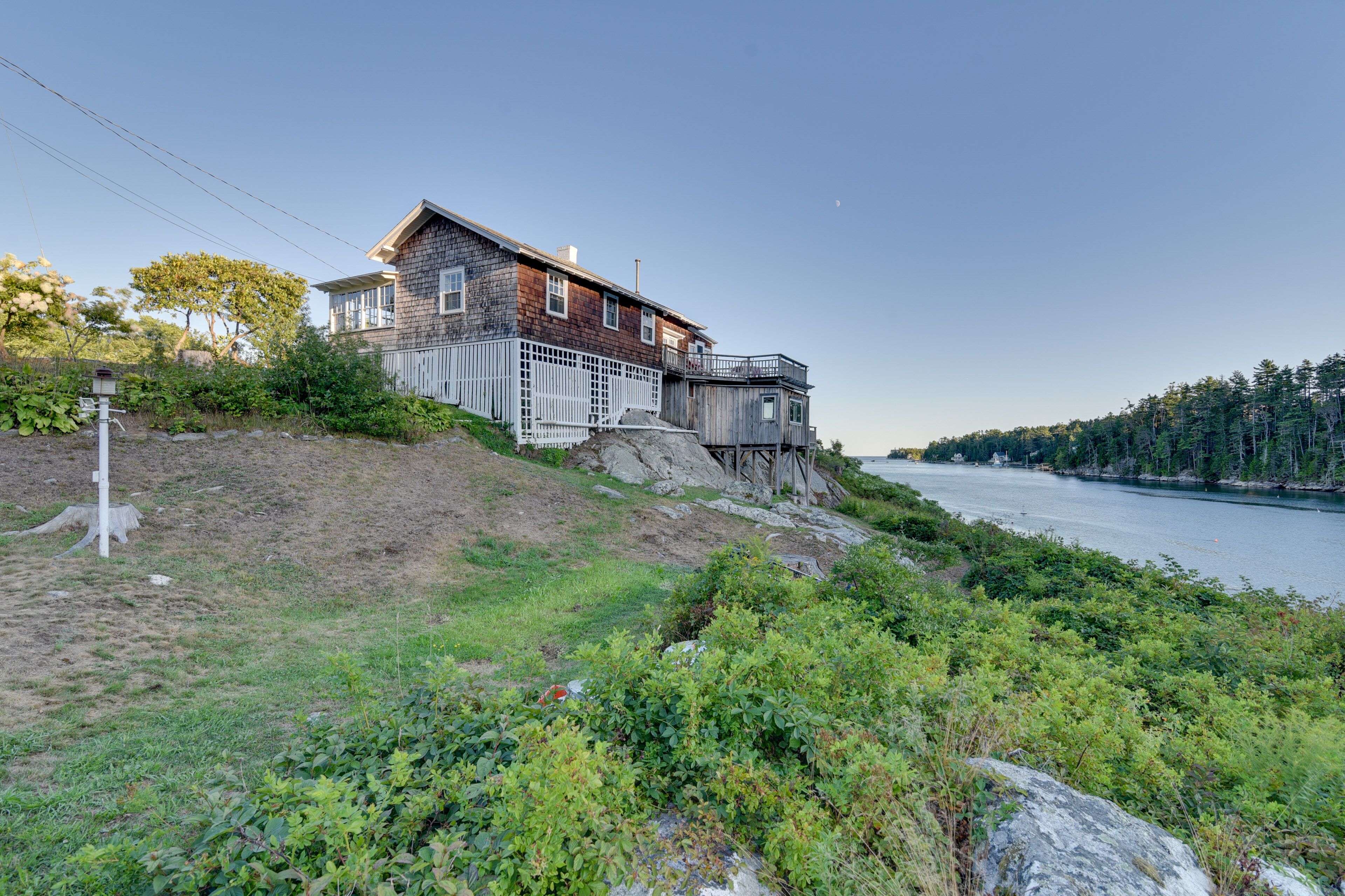 A two-story house with a wooden deck and a small cabin-like structure on a hillside overlooking a body of water.