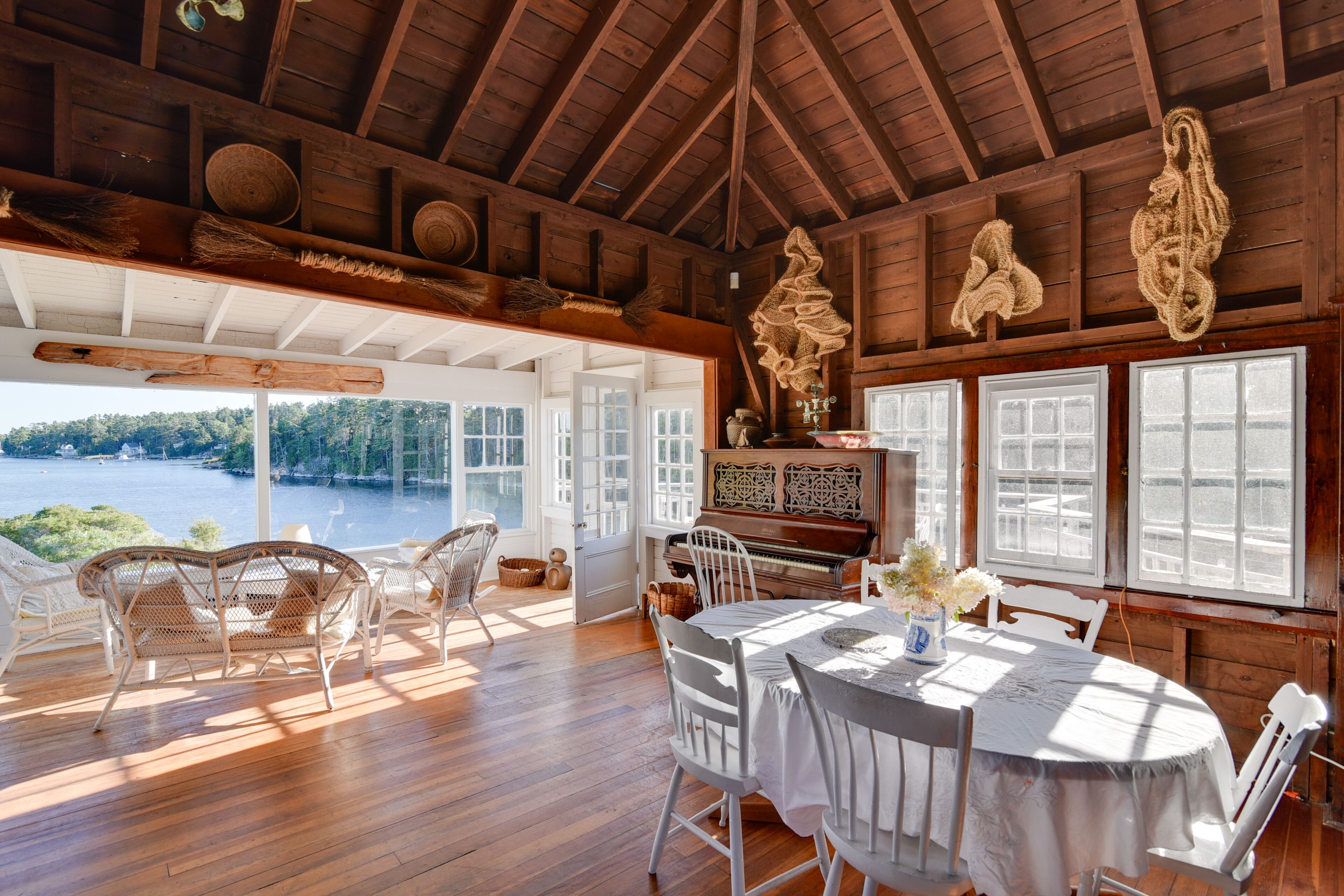 A wooden interior with a piano, wicker chairs, a dining table, and large windows overlooking a body of water.